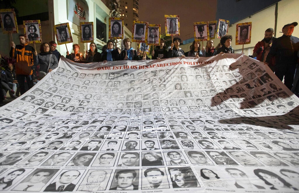 Manifestantes seguram um mural com fotos de pessoas mortas durante a ditadura no Brasil do lado de fora da delegacia que costumava ser centro de tortura da ditadura (DOPS), durante um protesto contra a remoção feita pelo presidente Jair Bolsonaro de membros de uma comissão que investigava crimes cometidos durante a ditadura brasileira em São Paulo. Segunda-feira, 5 de agosto de 2019