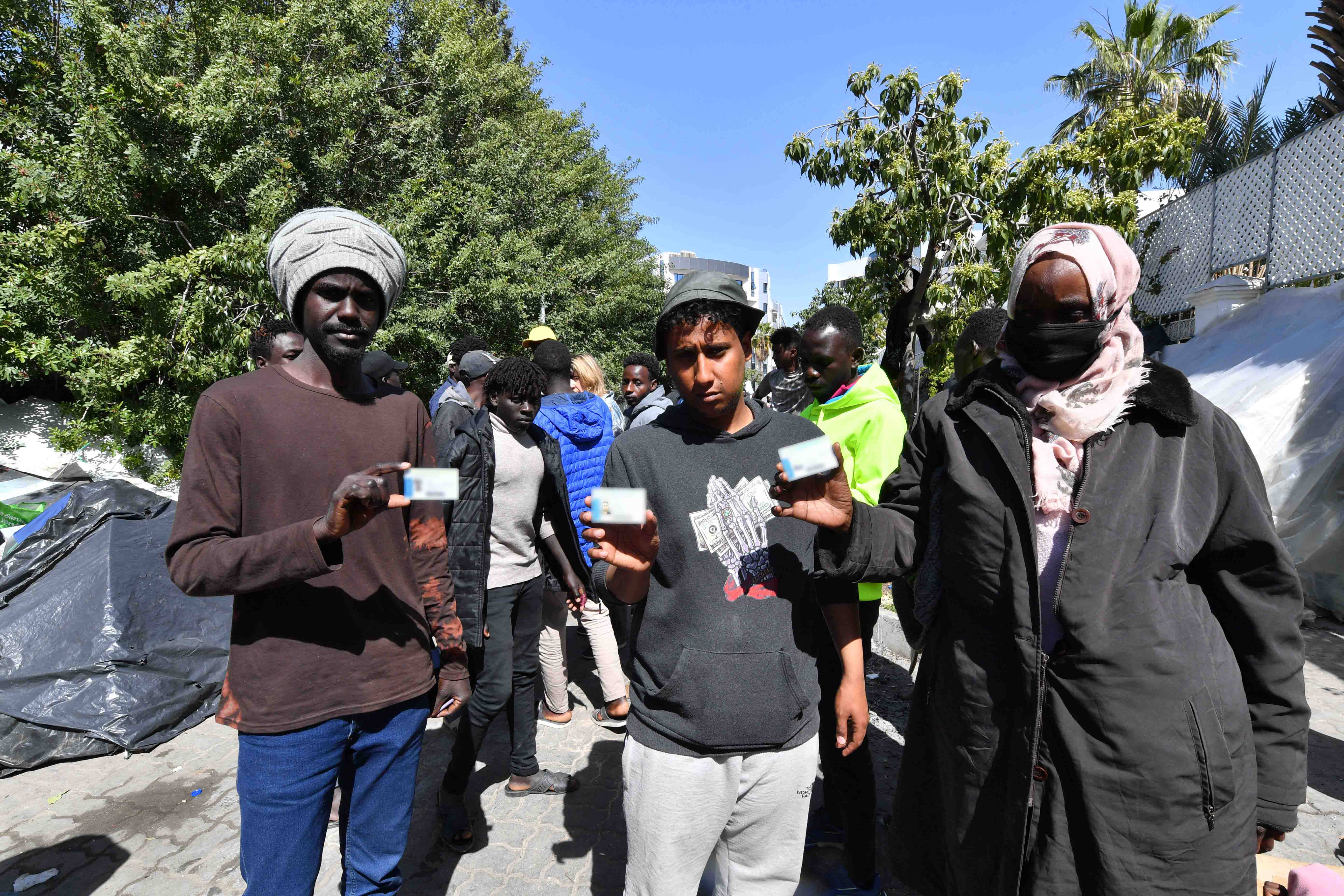 أشخاص سودانيون ويمنيون يعرضون بطاقات اللاجئين أو طالبي اللجوء الخاصة بهم في 12 أبريل/نيسان 2023 أمام مكتب "المنظمة الدولية للهجرة" في تونس العاصمة، بعد استخدام الشرطة التونسية في 11 أبريل/نيسان القوة والغاز المسيل للدموع لتفريق اللاجئين وطالبي اللجوء والمهاجرين وتفكيك مخيمهم المؤقت أمام المكتب القريب لمفوضية الأمم المتحدة للاجئين في تونس.  ©2023 فتحي بلعيد/أ ف ب عبر غيتي إيمجز 