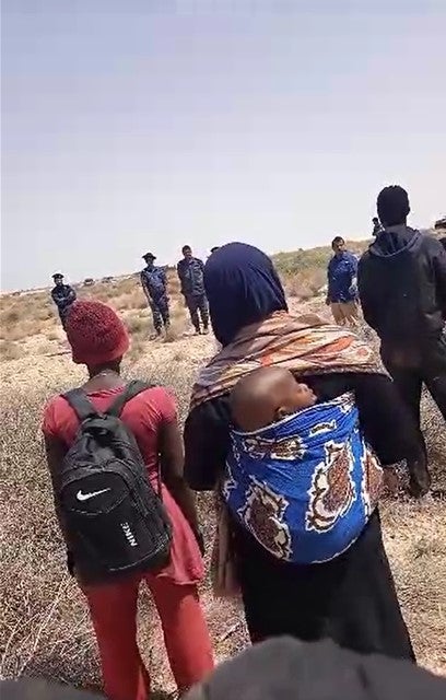 مجموعة من المهاجرين وطالبي اللجوء السود من جنسيات أفريقية متعددة، بينهم امرأة وطفلها، تقطّعت بهم السبل في الصحراء لعدة أيام بعد طردهم من تونس، يقفون في المنطقة العازلة على الحدود التونسية-الليبية مقابل طاقم إخباري لقناة "الجزيرة" وجنود ليبيين، 11 يوليو/تموز 2023. © 2023 خاص