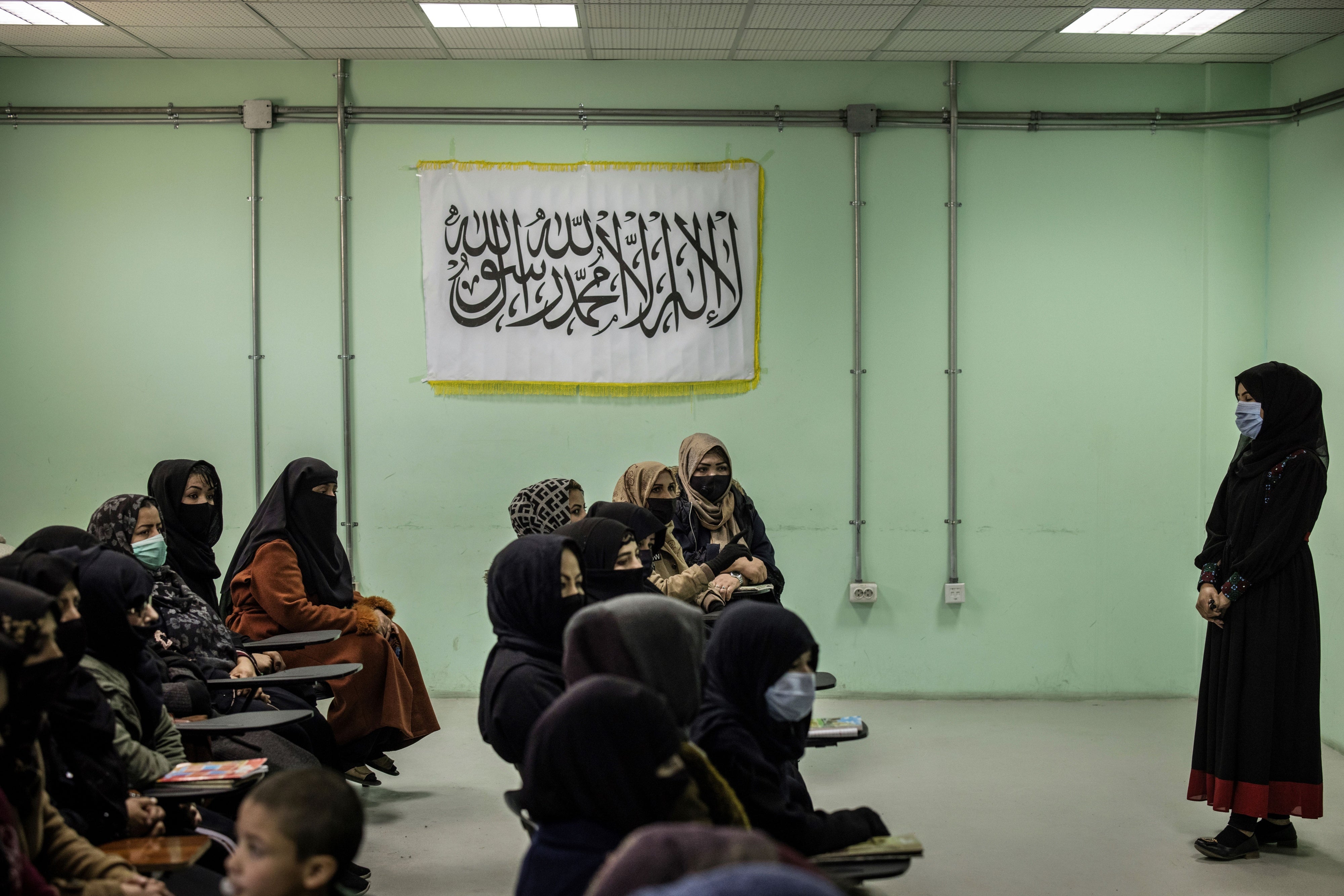 Women seated in a classroom with the flag of the Taliban hanging on the wall