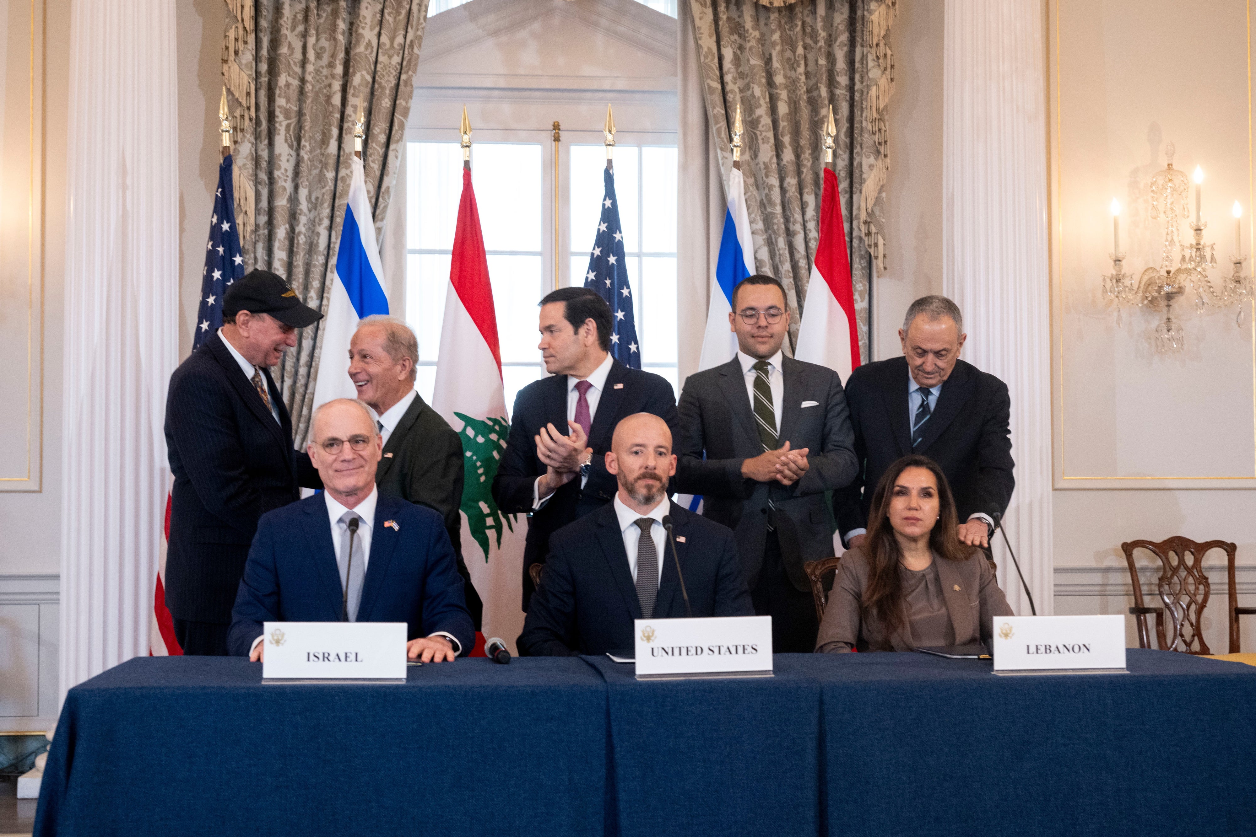 Three people sit at a table with the plaque cards for the US, Israel, and Lebanon while others behind them clap.