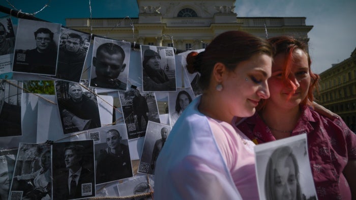 Two women hold a photo of a person in front of a fence of other photos of prisoners