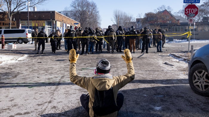 A protester sits on the street with his arms up in front of federal agents and Minneapolis Police