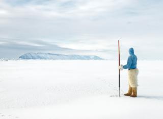Photograph showing an indigenous man holding a spear and standing on a sea of ice looking out at snow capped mountains.