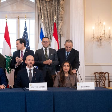 Three people sit at a table with the plaque cards for the US, Israel, and Lebanon while others behind them clap.