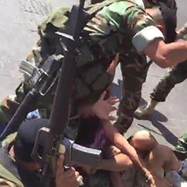 A group of armed men in army uniforms beat two people lying on the ground, one trying to shield the other.